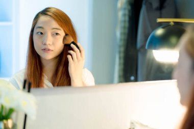 Genç Asyalı bir kadın makyaj fırçasıyla evde aynanın karşısına geçiyor, rahat, aydınlık bir soyunma odasında güzellik rutinine odaklanıyor..