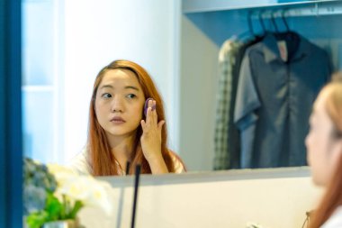 Genç Asyalı bir kadın aynanın karşısına yastıklı puf kullanarak makyaj yapıyor, güne odaklanarak ve özenle hazırlanıyor şık, modern bir yatak odasında..