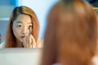 Genç Asyalı bir kadın evde aynanın önünde bir fırçayla göz makyajı yapıyor, sakin ve rahat bir şekilde güzellik rutinine odaklanıyor..