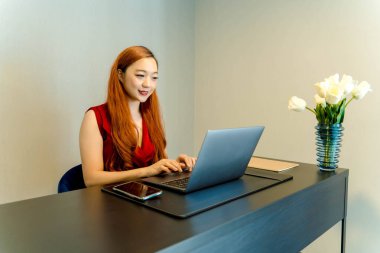 Asyalı bir kadın, elinde çiçek vazosuyla modern bir masada oturmuş, minimalist çalışma alanı düzenine odaklanmış ve gülümsüyor..