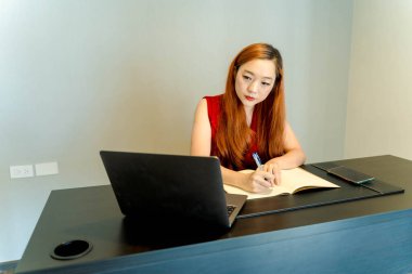 Asyalı bir kadın evde çalışırken not defteri yazıyor. Bilgisayarı ve akıllı telefonu olan siyah bir masaya odaklanmış ve meşgul. Minimalist modern çalışma alanı..