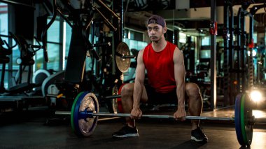 Kırmızı kolsuz gömlek giyen genç Asyalı adam halterle halterle modern spor salonunda ağırlık kaldırıyor. Gücünü, formda olduğunu ve düzenli egzersiz yaptığını gösteriyor.