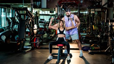 Spor salonunda halterli omuz egzersizi yaparak kadın müşteriye rehberlik eden özel antrenör, uygun forma ve kuvvet eğitim tekniğine odaklanıyor.