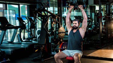 Spor salonunda otururken dambılları kaldıran sakallı bir adam, kas geliştirme ve vücut geliştirme eğitimine ve sağlıklı yaşam tarzına odaklanmış..