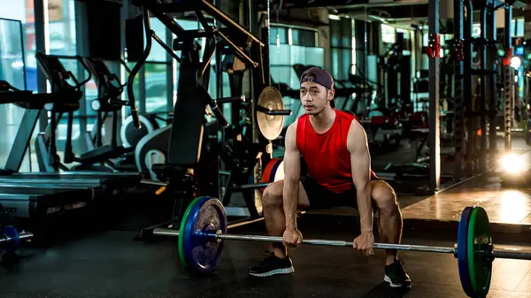Kırmızı kolsuz gömlek giyen genç Asyalı adam halterle halterle modern spor salonunda ağırlık kaldırıyor. Gücünü, formda olduğunu ve düzenli egzersiz yaptığını gösteriyor.