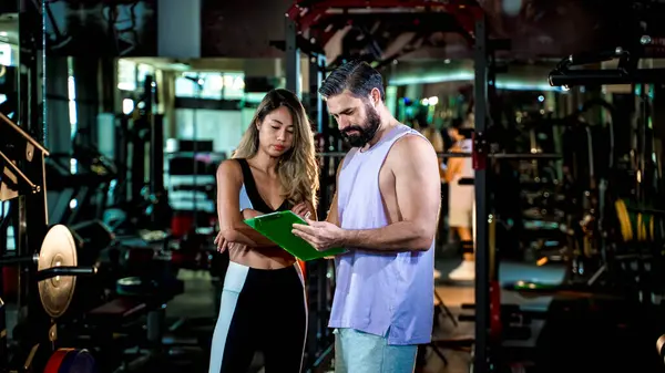 Beden eğitimi eğitmeni, modern spor salonunda bayan müşteriyle kişisel çalışma planını gözden geçiriyor. Koçluk, planlama ve başarı için fitness hedef oturumu.