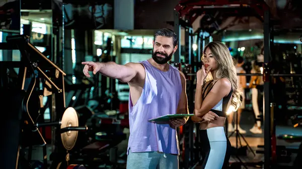 Spor salonunda bayan müşteriyle egzersiz stratejisini tartışan kişisel antrenör, elinde panoyla fitness hedeflerini planlarken ekipmanlara işaret ediyor.
