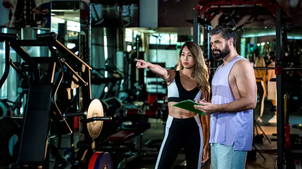 Spor salonundaki erkek müşteriye antrenman planının yönünü belirten kadın fitness koçu, kişisel antrenman sırasında attığı adımları açıklarken elinde bir not panosu tutuyor.