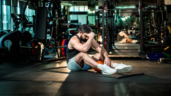 Sıhhiye topuyla yapılan yoğun egzersizden sonra spor salonunda oturan yorgun adam tükenmişliği, yorgunluğu ve fiziksel kararlılığı sembolize ediyor..
