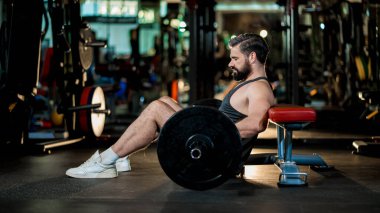 Siyah kolsuz tişörtlü güçlü adam kalça itme egzersizine hazırlanıyor. Spor salonunda halterli halter, kalça ve alt vücut çalışmalarına odaklanmış.