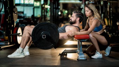 Spor salonundaki ağırlık kaldırma egzersizi sırasında erkek sporcuya yardımcı olan kadın kişisel antrenör form, güç ve düşük vücut gücüne odaklanıyor.