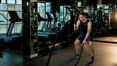 Odaklanmış genç adam modern bir spor salonunda güçlü ip dalgaları sergiliyor. Güç gösterisi, kondisyon ve tam vücut dinamik çalışma yoğunluğu.