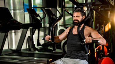 Modern spor salonunda göğüs pres makinesi üzerinde çalışan siyah kolsuz tişört ve gri şort giyen, vücudunun üst kısmını yoğun bir şekilde güçlendiren odaklanmış sakallı bir adam.