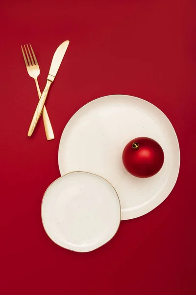 Elegant table setting with two plates, gold silverware and bright red christmas bauble on bold red background. Minimal celebration concept.