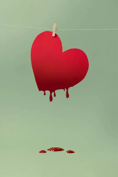 Ketchup dripping from a red heart hanging on a string with sage green background. Creative love concept.