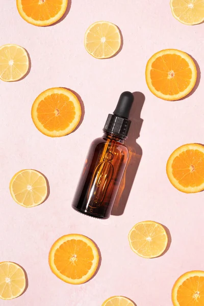 Kahverengi cam kozmetik düşürücü şişeli canlı C vitamini serumu konsepti ve pastel pembe arka planda limon ve portakal dilimleri. Doğal parlak cilt tasarımı.