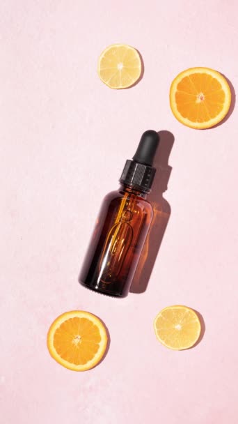 茶色のガラス化粧品ドロッパーボトルとパステルピンクの背景にレモンとオレンジのスライスと鮮やかな新鮮なビタミンC血清 自然な明るいスキンケアの概念 — ストック動画