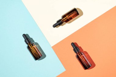 Renkli sarı, turuncu ve mavi arka planda üç kahverengi cam kozmetik serumu veya yağ damlası şişesi. En az cilt bakım ürünü tasarımı.