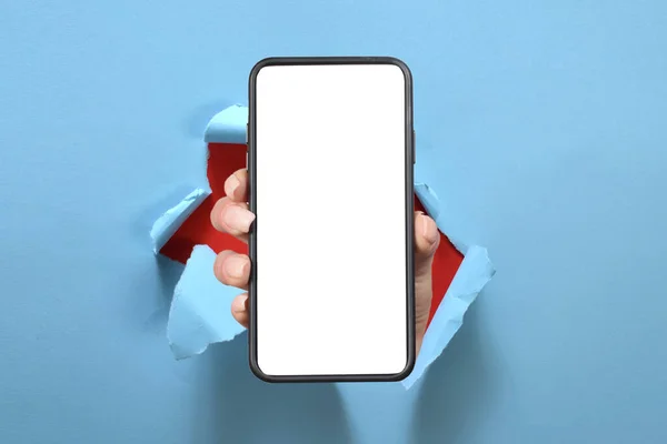 青い紙のシートを突破するカメラの近くに白い空の画面のデバイスを示す手のスマートフォンを保持 — ストック写真