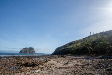 A scenic view of the coastline at Diguisit, Quirino, Philippines, featuring a rocky beach leading to the sea with a prominent island in the distance. Lush green vegetation covers the hillside on the right, contrasting with the rocky foreground. This  clipart