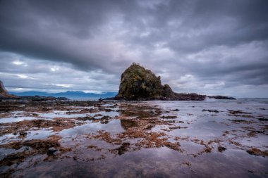 Diguisit 'in dramatik bir sahnesi, Aurora' nın kumlu bir sahilde, hızlı hareket eden, karamsar bulutlarla dolu bir gökyüzünün altında iki engebeli kaya oluşumu. Sakin su, gökyüzünün zengin tonlarını yansıtarak çarpıcı ve atmosferik bir deniz manzarası yaratır..