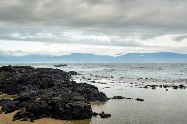 Diguisit 'in dramatik bir sahnesi, Aurora' nın kumlu bir sahilde, hızlı hareket eden, karamsar bulutlarla dolu bir gökyüzünün altında iki engebeli kaya oluşumu. Sakin su, gökyüzünün zengin tonlarını yansıtarak çarpıcı ve atmosferik bir deniz manzarası yaratır..