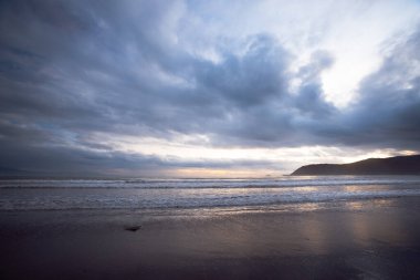 Alacakaranlıktaki bir kıyı şeridinin büyüleyici görüntüsü, koyu renkli bulutlarla dolu dramatik bir gökyüzü. Yumuşak, dağınık ışık bulut örtüsünü kırıyor, sakin okyanusu aydınlatıyor ve bir huzur ve sükunet hissi yaratıyor. Bu çağrışım yapıyor.