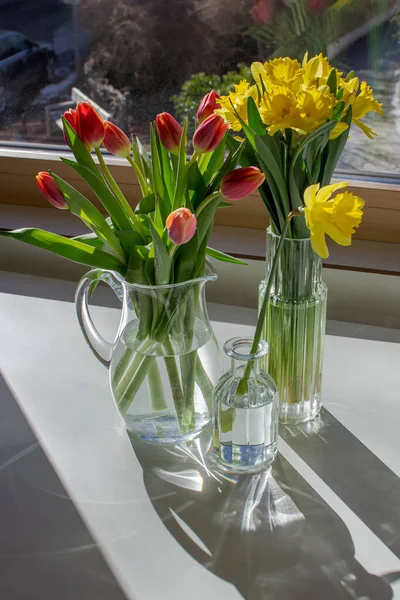 Three glass vases with tulips and daffodils on a white table in the sun
