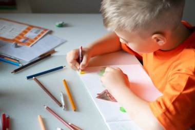 Turuncu tişörtlü bir çocuk beyaz bir çarşafa renkli kalemler çiziyor..