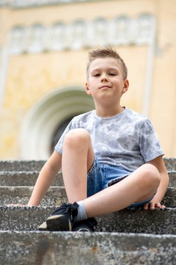 Sıradan giyinmiş bir çocuk parkın merdivenlerinde oturur, düşünceli gözleri kameraya bakar, sessiz bir yansımayı yakalar..