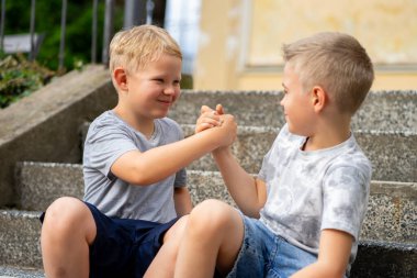 İki çocuk merdivenlerde bilek güreşi yaparken gülümsüyor, dostça bir yarışmanın ve sıcak bir yaz gününün tadını çıkarıyor..