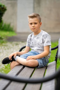 Genç bir çocuk ahşap bir bankta tek başına oturuyor, yüzünde sakin ve düşünceli bir ifadeyle doğrudan kameraya bakıyor..