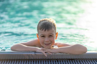 Gün batımında yüzdükten sonra açık havuzun kenarına tutunan gülümseyen çocuk.