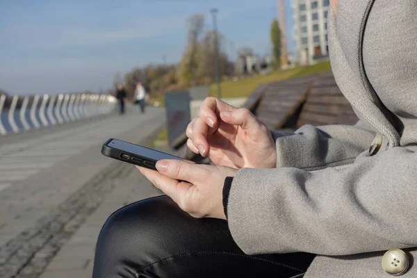 Kadın eli bankta otururken cep telefonu kullanıyor. Kız akıllı telefonu açık havada tutuyor. Gri montlu, fularlı bir kadın parkta bir bankta cep telefonu tutuyor.