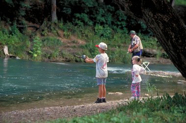 A little girl watches her big brother fish while their grandfather casts in the background at Roaring River State Park in Missouri. clipart