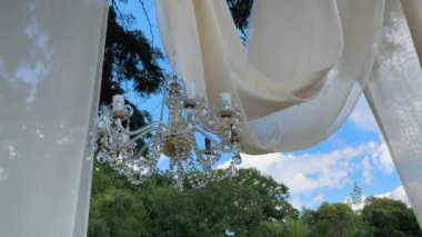 Elmas elmaslı kristal avize rüzgarda ve parlaklıkta pürüzsüzce döner. Bir avize ve beyaz tül rüzgârda savruluyor doğanın arka planına karşı. Yüksek kalite 4k görüntü