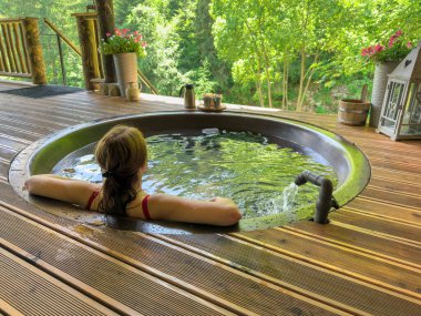 Kız geleneksel demir bir jakuzide dağ nehrinin arka planına karşı yıkanır. Şifalı bitkilerle su ateşte ısıtılır. Dağ tatil köyü spa ve eğlence. Yüksek kalite fotoğraf