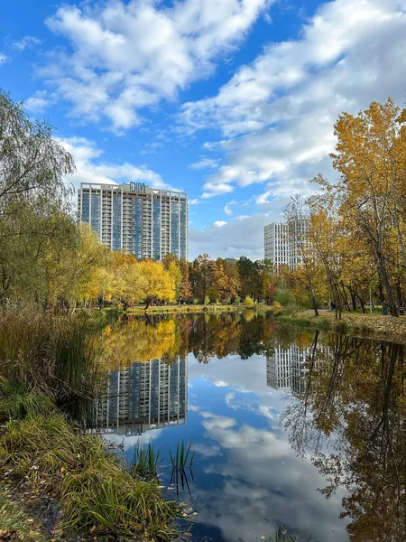 Beautiful autumn landscape of a city park in October. Multi-colored trees and multi-story buildings are reflected in the calm water of the lake. Vertical photo