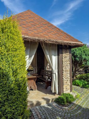 Güneşli bir günde Beyaz Perdeli Cozy Garden Gazebo. İyi Bakılmış Bahçe 'de Ahşap Benekli Zarif Açık Hava Pavyonu. Döşeme ve Dekoratif Taş işçiliğiyle Büyüleyici Arka Bahçe Çardağı. Yüksek