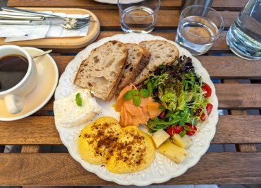 Açık bir kafede nefis bir kahvaltı, içinde Benedict yumurtası, somon balığı, taze salata ve ekmek bulunan cömert bir tabakla servis edilir. Dekoratif bir tabakta dengeli bir kahvaltı yemeğine yakın çekim. Yüksek kalite