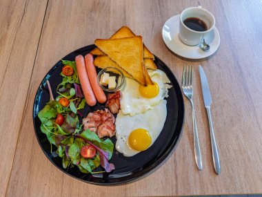 Sıcak ve eksiksiz bir kahvaltı tabağı, içinde güneşli yumurtalar, sosisler, çıtır pastırma ve tost var. Afiyet olsun ve mükemmel servis edilmiş kahvaltı. 
