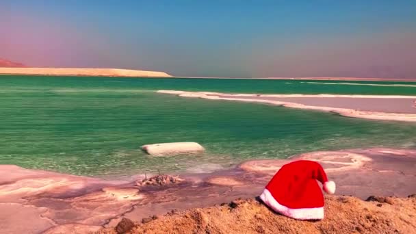 イスラエルの死海の岸にあるサンタの帽子 雪の代わりに白い塩 冬の夏 クリスマス休暇 夏への冬の旅行 熱帯の目的地 砂漠の新年の概念 — ストック動画