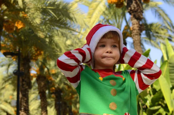 Positive funny kid in Santa Claus hat and elf costume in palm grove. Portrait of a child who holds his head, covers his ears with his hands. Christmas travel, vacation at the resort in winter. Children\'s whims, surprise, joy, tired.