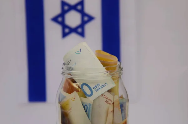 イスラエルの国旗を背景にイスラエルのシェケルとガラス瓶 個人貯蓄 銀行システム インフレ 国民通貨 — ストック写真