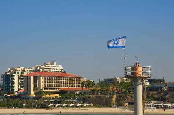 stock image Lighthouse in Herzliya marina. Flag of Israel in the wind. The beach and surfers are in the background. Panoramic view of the Israeli respectable resort
