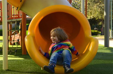 Çocuk parkında renkli bir kazak giyen sevimli bir çocuk. Çocuk gülüyor, oynuyor, kaydırak kayıyor. Çocuk tatili, eğlence parkı, tatiller, aile tatili, anaokulu kavramı..