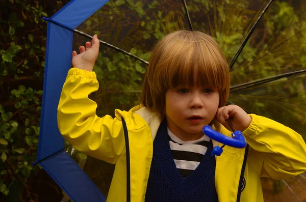 Cute positive boy in a yellow raincoat with a transparent umbrella. Cloudy weather, rain, the child walks on the street. Portrait of a child in the rain