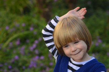 Sarı uzun saçlı, gülümseyen, gülümseyen, çekici bir çocuğun portresi. Arka planda çimenler ve mor çiçekler var. Anneler günü kartı, bahar günü kartı, anaokulu reklamı.