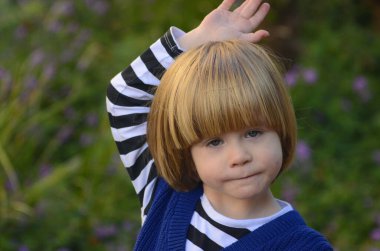 Sarı uzun saçlı, gülümseyen, gülümseyen, çekici bir çocuğun portresi. Arka planda çimenler ve mor çiçekler var. Anneler günü kartı, bahar günü kartı, anaokulu reklamı.