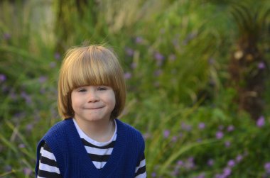 Sarı uzun saçlı, gülümseyen, gülümseyen, çekici bir çocuğun portresi. Arka planda çimenler ve mor çiçekler var. Anneler günü kartı, bahar günü kartı, anaokulu reklamı.